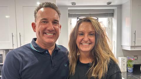 A man with short black hair is looking at the camera smiling. He is wearing a blue polo neck top and is standing in a kitchen. Next to him is a woman with long brown/blonde hair and glasses on her head. She is smiling at the camera. 