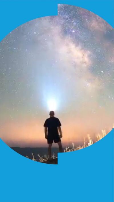 The outline of a man facing away from the camera wears a head torch and looks out into a beautiful night sky. There is an array of stars and the colours of the sky fade from blue to orange.
