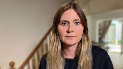 Polly has long blond hair. She stands in a house with the staircase behind her wearing a black jumper. 