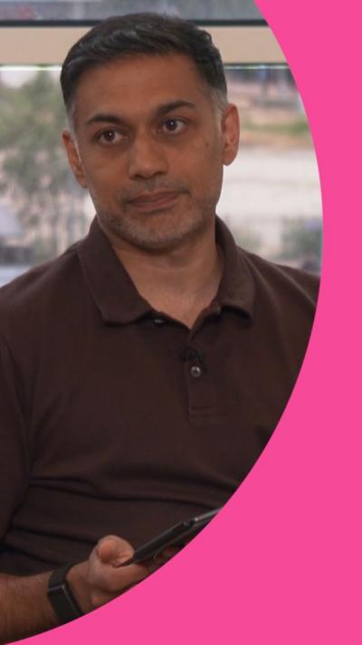 A man with short dark hair looks into the camera. He is holding a tablet and wearing a brown T-shirt with a button-up collar.