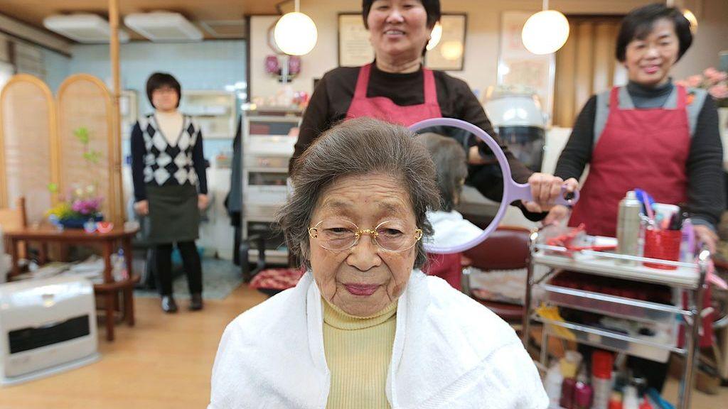 A 100-year-old Japanese woman wearing a butter yellow turtleneck, white jacket and rimless glasses at a hair salon with a hairdresser holding up a mirror behind her head