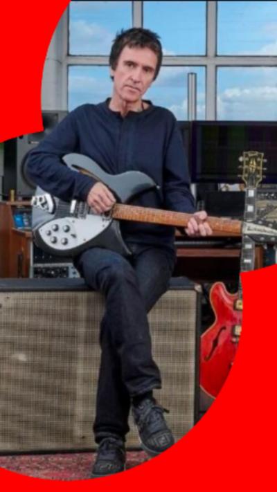 Johnny Marr sat on an amp playing a guitar with other guitars surrounding him.