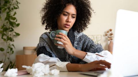A woman wrapped in a blanket and holding a hot drink sits at a table with a laptop on it. The table has scrunched-up tissues on it as well. 