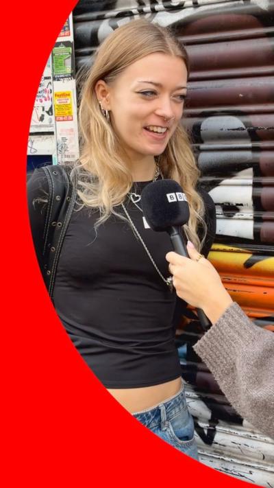 A young woman with nose piercings, blue jeans, and a black top speaking into a BBC branded microphone, looking like she's in conversation 
