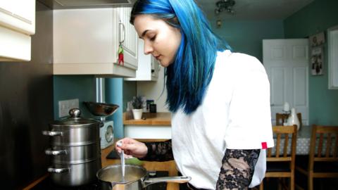 A young woman with dyed blue hair and white top stirs a pot of boiling water on a stove in a kitchen