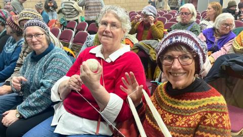 A group of female knitters are sitting in chairs in a large hall. Some of them are wearing woollen hats, while the two women nearest the camera are working with a ball of wool.
