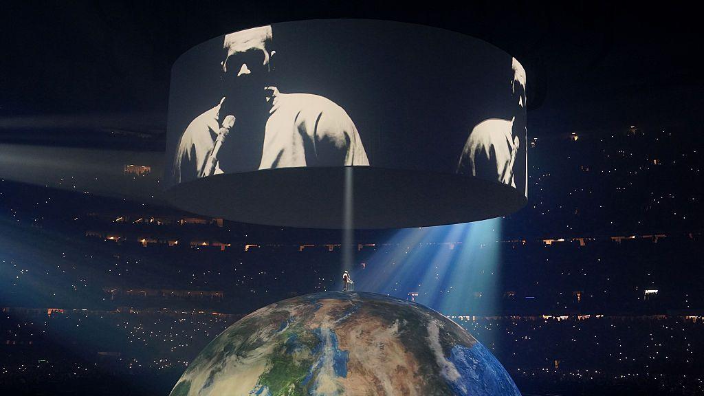Kanye West performing in New Orleans. Ye stands atop an enormous globe, a spotlight beaming down on him. Above he is projected on screens to a crowd that surrounds him. 