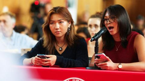 Two female students with Cornell University’s ACLU, a student organization that advocates for civil rights, address the student assembly on Sexual Violence held at Cornell University. They are speaking in to microphones at a red coloured table