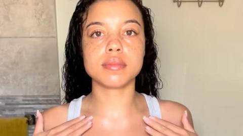 A head and shoulders photo of Abbey Alexandra with long dark curly hair facing the camera wearing a white vest top with her fingertips touching her collarbones