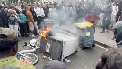 A large commercial bin is seen on fire on a road, surrounded by teenagers