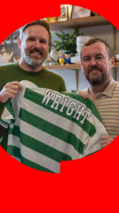 An image of two middle aged men holding a green and white Celtic football strip in a kitchen. They both have beards and short dark hair.