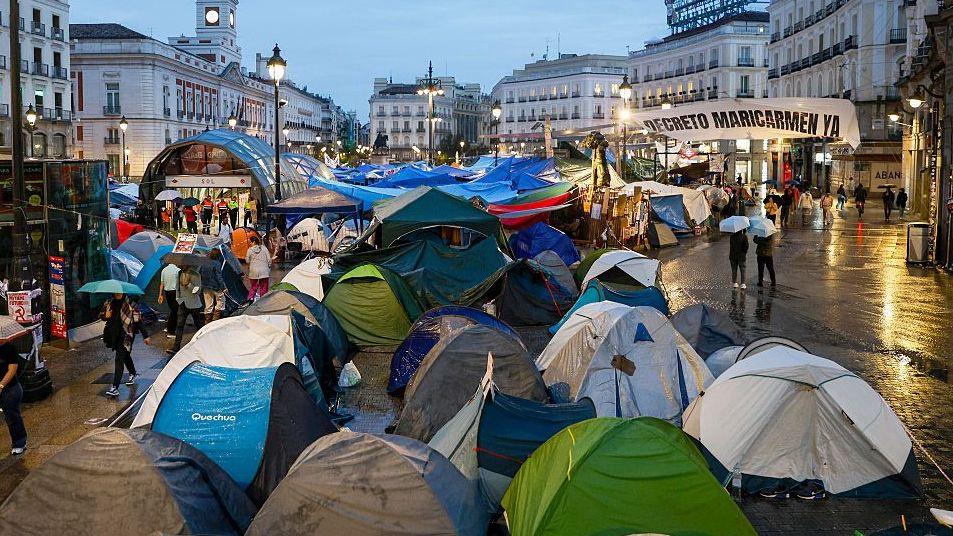 About a dozen small tents are pitched in a concrete square in the centre of Madrid. It is raining and people walk around the tens with umbrellas. 