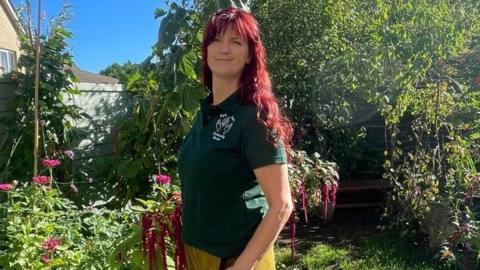 Stefi Robertshaw is pictured standing in a garden on a clear and sunny day. She has long red hair which is wavy and she is wearing a dark green T-shirt with a white logo. Green plants, trees and pink flowers can be seen in the background.