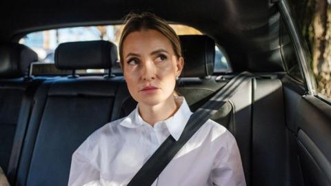 A woman wearing a white shirt sits in the back of a car. She is looking out the window and is waring a seatbelt. She doesn't look happy.