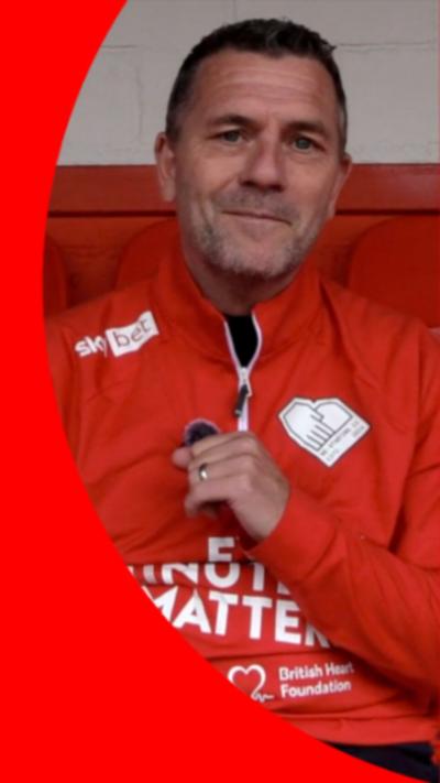 Man with dark brown hair wearing a red track suit top seated