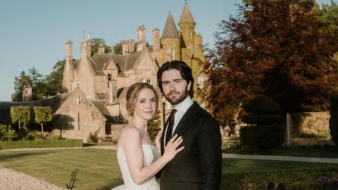 Savannah who is blonde, fair skinned and wearing a white wedding dress stands next to her husband Hayden, who has dark hair and a beard and wearing a black suit. They stand in front of a Scottish Castle with the sunshining. 