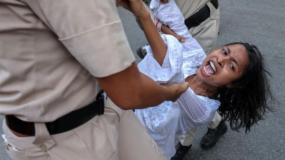 Two uniformed men drag a woman she appears to be shouting. She is wearing a white shirt and has long, black hair. 