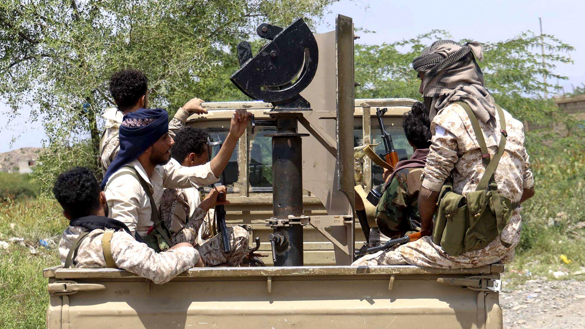 Several people ride in the back of a pick-up truck carrying mounted equipment along an unpaved road lined with trees and scrub