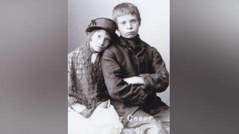 An old black and white photo shows a girl and a boy sitting next to each other. The girl is leaning her head on her brother's shoulder. She is wearing a dark coloured hat and a shawl and dress or skirt. Her brother is wearing a dark coloured jacket and trousers and a scarf. He has his arms folded. Both are looking at the camera. Some white writing which is barely legible but whcih ends with the word 'group' can be seen on the bottom of the photo.