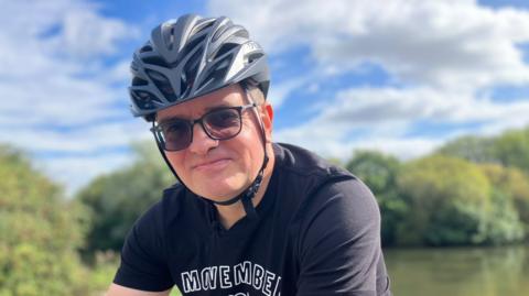 Ben Lidgey is wearing a grey cycling helmet and glasses. He is sat on a bicycle. Ben is wearing a black T-shirt with the visible text “MOVEMBER AMBASSADOR.” Trees and greenery are in the background with a partly cloudy sky. 