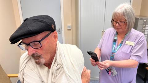 A man in his seventies who is wearing a black flat cap and glasses is lifting up his shirt to reveal his back.  A healthcare assistant is taking pictures of his back using a device that looks like a mobile phone.