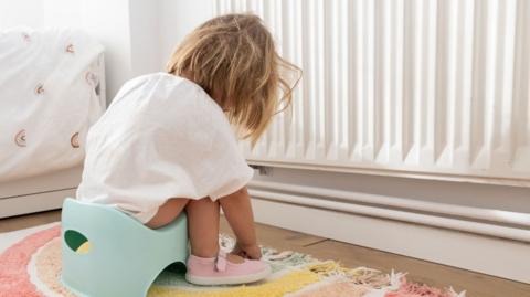 Child sitting on a small potty in a bedroom, viewed from behind. The child is wearing an oversized white top and pink shoes, seated on a pastel potty on a colourful rug beside a radiator.