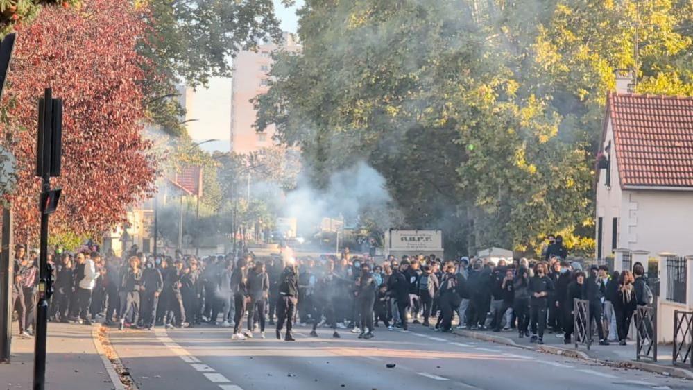 A large crowd of students gathers on a tree-lined street as smoke drifts across the road, with several individuals standing in the foreground