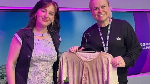 Two women are smiling as they stand in the Radio Lancashire studio. The woman on the right is holding an off-white aged short sleeved top.