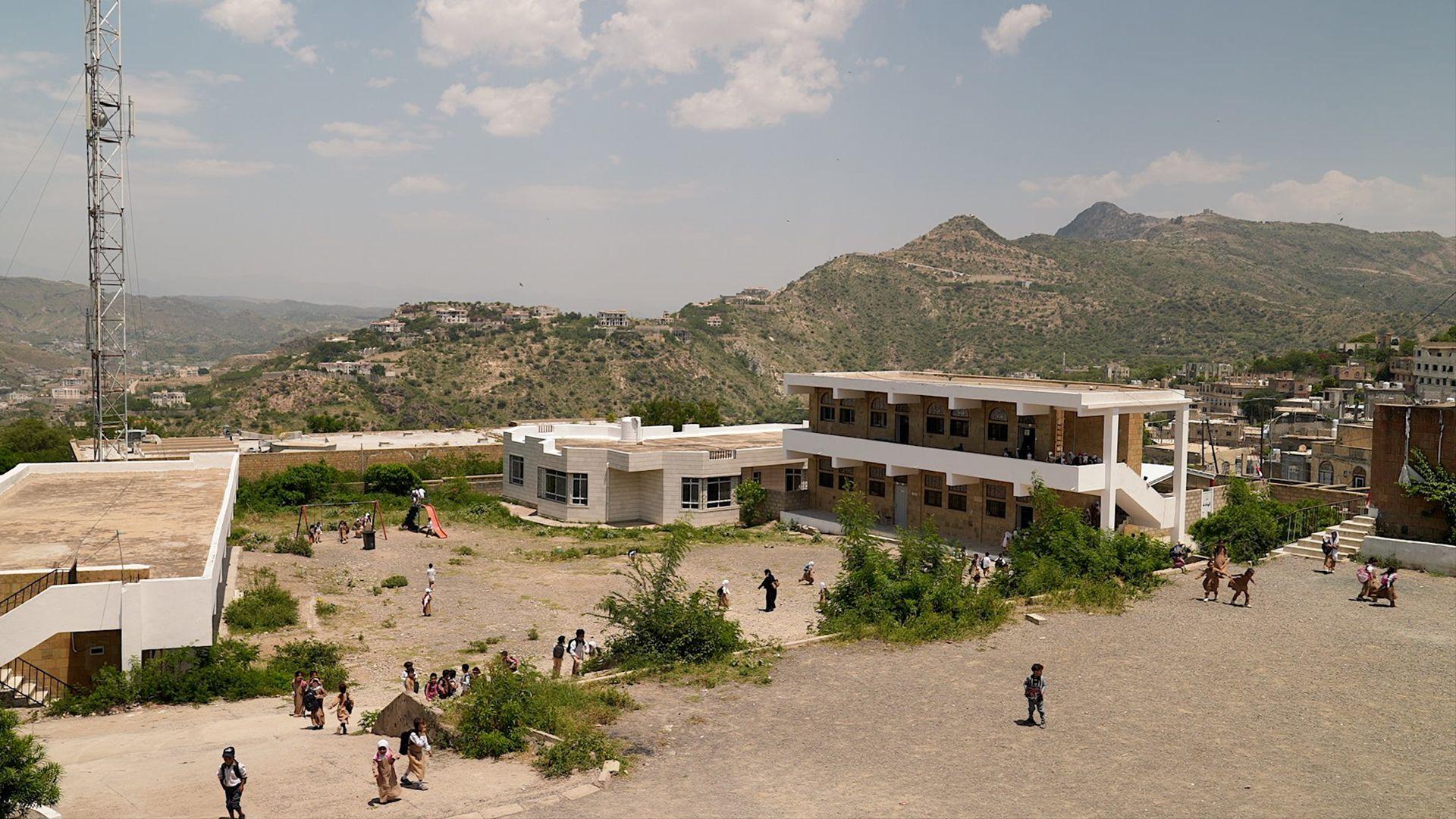 A school complex made up of low, white buildings positioned around a yard. There are about 20 people dotted around the foreground, and there are mountains in the background. 