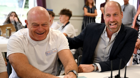 Ross Kemp and Prince William laugh as they sit by a table inside a room full of people.
