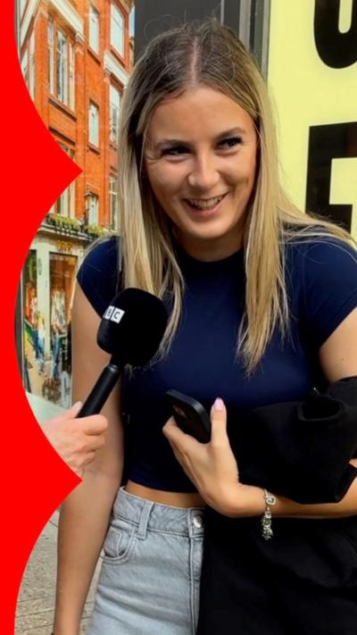 A woman stands in the street, wearing a navy top and smiling in front of a mic.