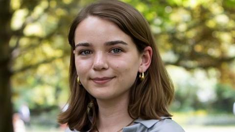 A woman with short brown hair in a bob smiling, she is wearing gold hoop earrings and a blue blouse. The background is blurred but features a large green tree. 