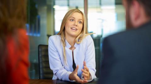Woman sits at a table in an office being interviewed by a man and a woman whose blurred backs are in the foreground.