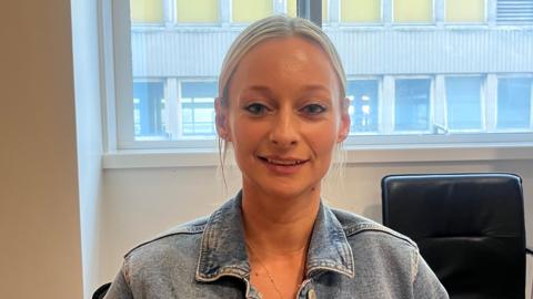 A blonde woman with her hair tied back. She wears a denim jacket 