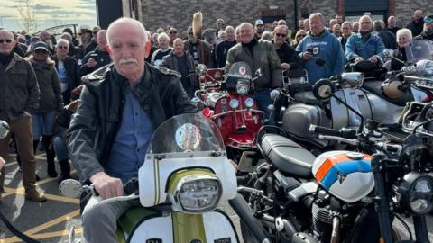 A man with white hair and moustache sits on a vintage-style scooter in the foreground, surrounded by dozens of people and rows of motorcycles at an outdoor gathering. Several scooters and motorbikes, including red, silver and blue models, are parked closely together, with a crowd standing behind them in a car park on a bright day.