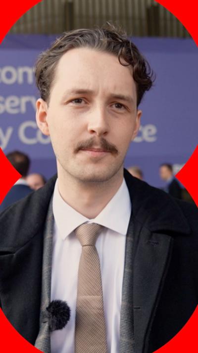 A young man talking to the camera at the Tory Party Conference. He has short dark hair and a moustache. He is wearing a dark blue jacket, white shirt and  a taupe tie.