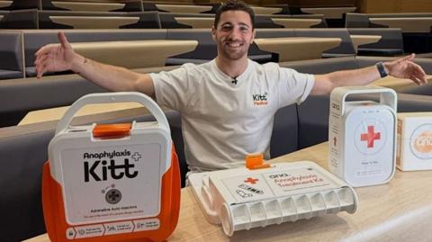 A man is sat with his arms outstretched, with some medical kits on display in front of him. 