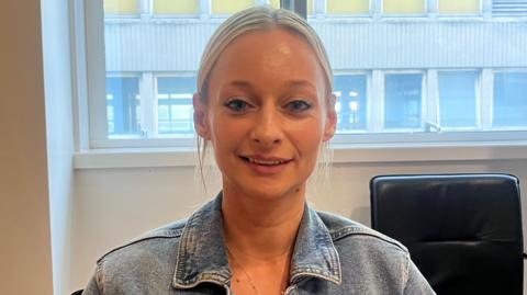 A blonde woman with her hair tied back. She wears a denim jacket 