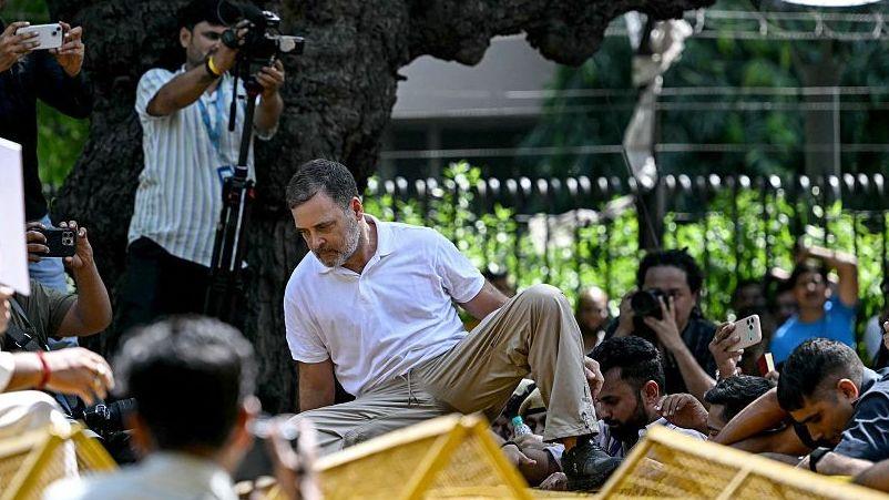 Opposition MP Rahul Gandhi of the Congress party, sporting a white polo T-shirt and khaki pants, climbs a police barricade. He is surrounded by security guards, supporters, police personnel and the media.