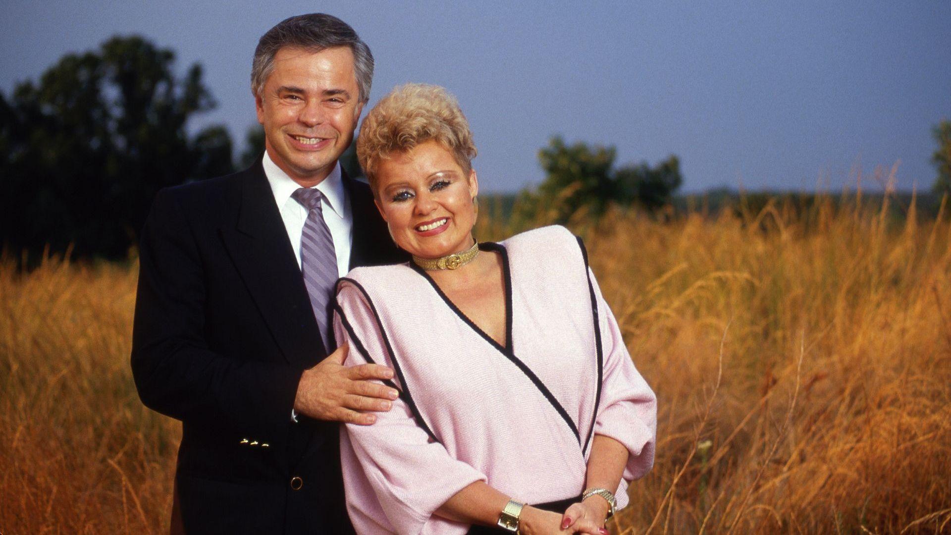 Jim and Tammy Faye Bakker on a hill overlooking their megachurch complex, Heritage USA in August 1987. Jim Bakker is in a suit smiling and holding Tammy Faye who is in a pink shirt. 