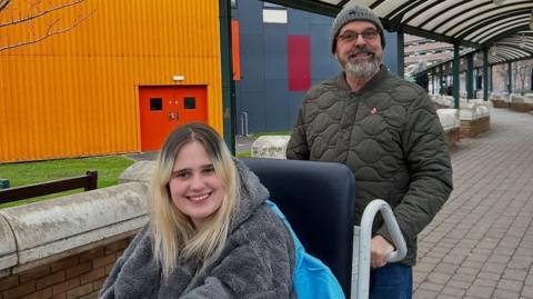 Phoebe Tesoriere is smiling at the camera and wearing a grey fleece jacket. She has long blonde hair. She is in a wheelchair.