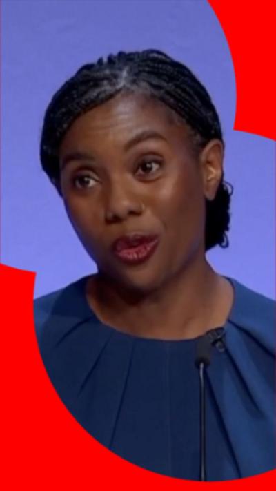 Kemi Badenoch is giving a speech. She is wearing a blue top with a round neckline and pleats over the chest.