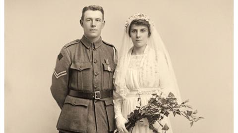 Sepia-toned studio wedding portrait showing two adults standing side by side against a plain backdrop. One person wears a military uniform with medals and insignia, while the other wears a light-coloured wedding dress, veil and gloves, holding a bouquet of foliage. Both face the camera in a full-length formal pose.