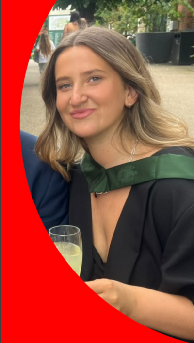 A young woman holds a glass of prosecco and is wearing a graduation gown. She is in her 20s and is smiling at the camera.