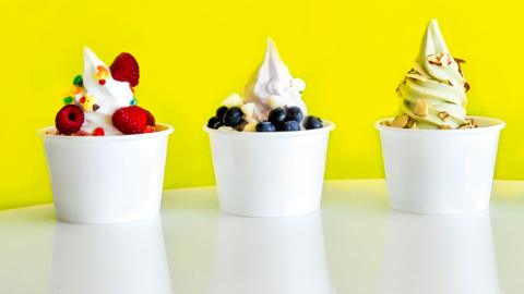 Three white tubs of frozen yoghurt, decorated with fruit and nuts, against a bright yellow background