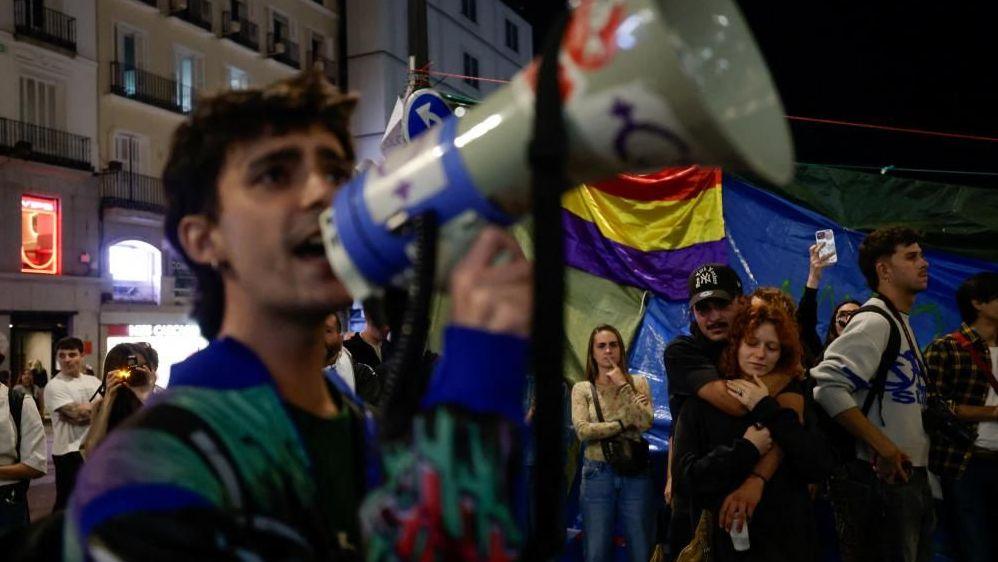 A demonstrator speaks into a megaphone in Puerta del Sol, Madrid. Two protesters are stood behind him hugging. 