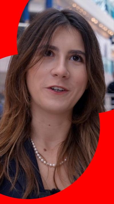 A young woman with long brown hair talks to the camera. She is wearing a dark-coloured top.