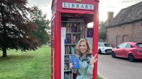 Sally Phillips leans against a bright red K2 phone box which has been transformed into a mini library. She is holding a copy of Winnie-the-Pooh from Alan Alexander Milne while smiling. The phone box is stacked with an assortment of free books, organised across various shelves.