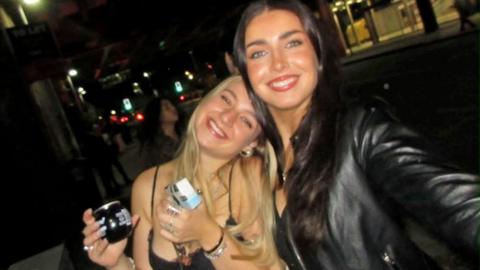 There are two girls in a dark bar smiling at the camera . On the left a blonde girl is wearing a strappy top and holding a drink. She leans into her friend on the left who is holding the camera to take a selfie of the two of them. The girl on the right has long brown hair and is wearing a black leather jacket.
