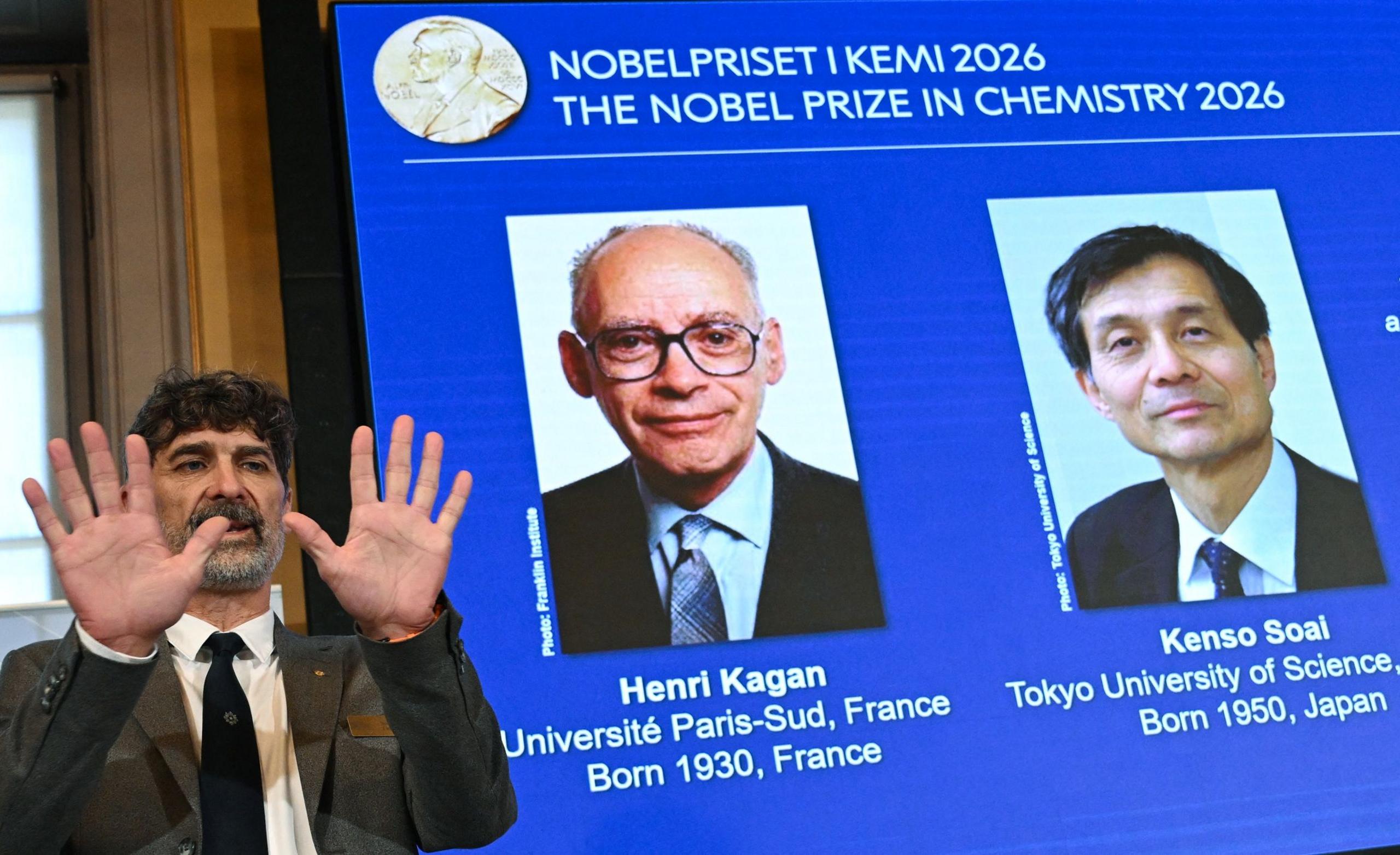 A man in a grey suit and black tie stands to the left of the picture and holds up both hands. There is a screen behind him that is titled in Swedish and English "The Nobel Prize in Chemistry 2026" below that are two pictures on the left a man in a black suit and blue tie - Henri Kagan - and to the right a man also in a black suit and blue tie - Kenso Soai. 